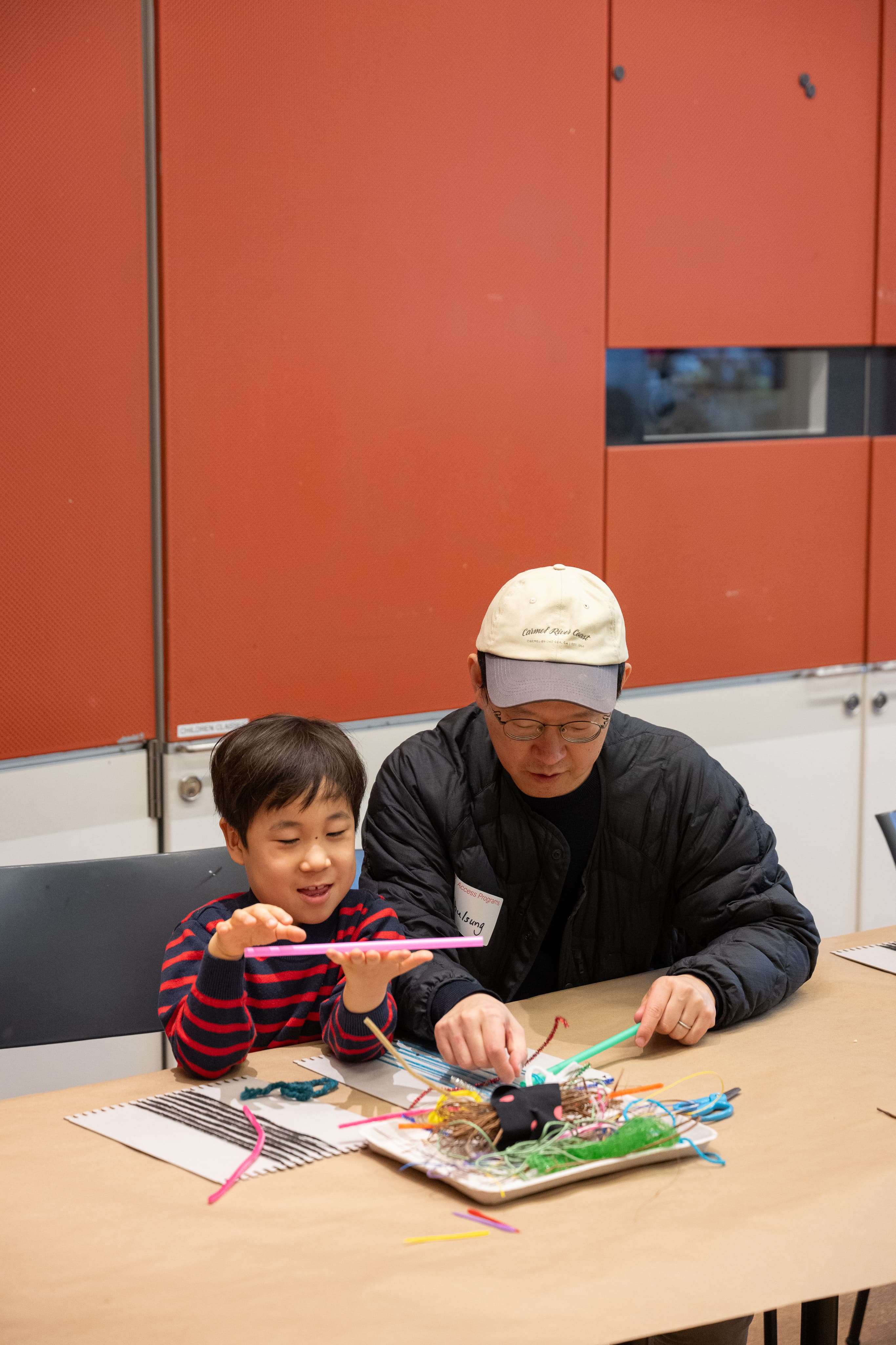 Two visitors sit at a table in a studio classroom at The Met. They are working together on a weaving project with pipe cleaners, fabric scraps, yarn, and small looms.