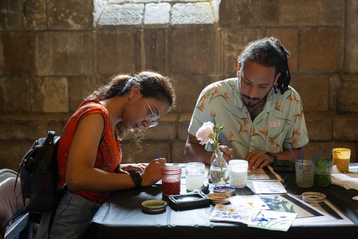 Two people paint while sitting at a table inside The Met Cloisters. The person on the left has glasses, an orange tank top, and dark hair in a ponytail. The person on the right is wearing a light blue shirt with orange flowers. On the table there is a flower in a vase, jars with water and paintbrushes, and floral paintings. Image: Photo by Filip Wolak