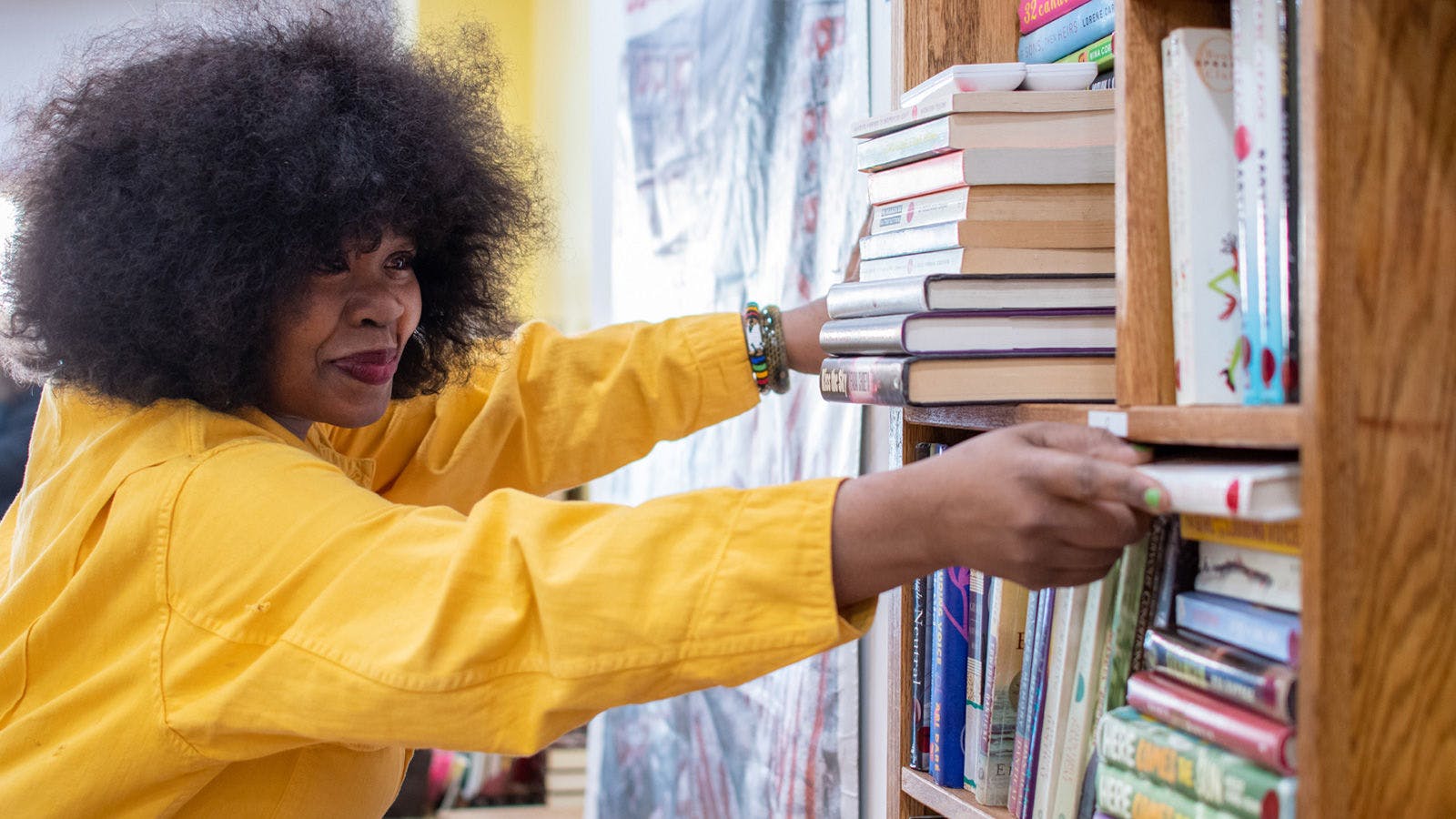 At left, a smiling woman in a yellow jacket reaches out with both hands towards a wooden bookshelf at right to retrieve a book. The shelves hold books both horizontally and vertically.