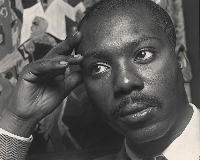 A black-and-white photo of Jacob Lawrence resting his head in his hand in front of one of his paintings