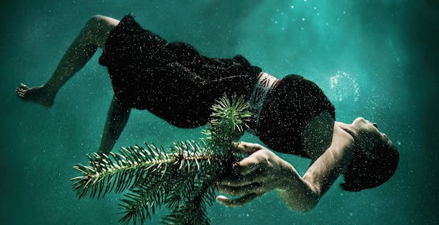 Water Memory Series photograph by Cara Romero. A woman wearing indigenous clothing floats underwater holding a sprig of fir needles.