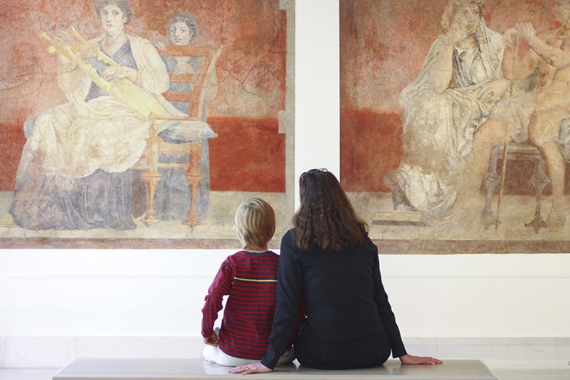 A child and an adult seated next on a bench admiring a work of art. They are photographed from behind.